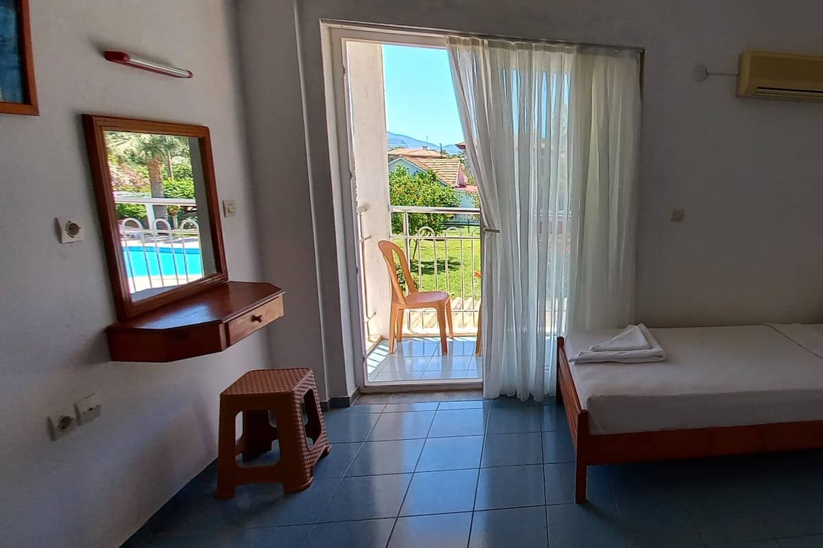Un dormitorio con cama, un taburete pequeño, una mesita de noche, un ventanal con vista a un balcón y un equipo de aire acondicionado.