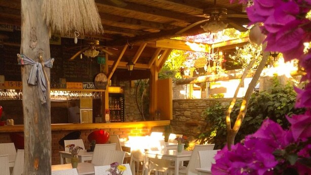 An outdoor dining area with white wicker tables and chairs, a stone floor, and a bar area with a rustic wooden ceiling. - Eski Datca Evleri (Datca)