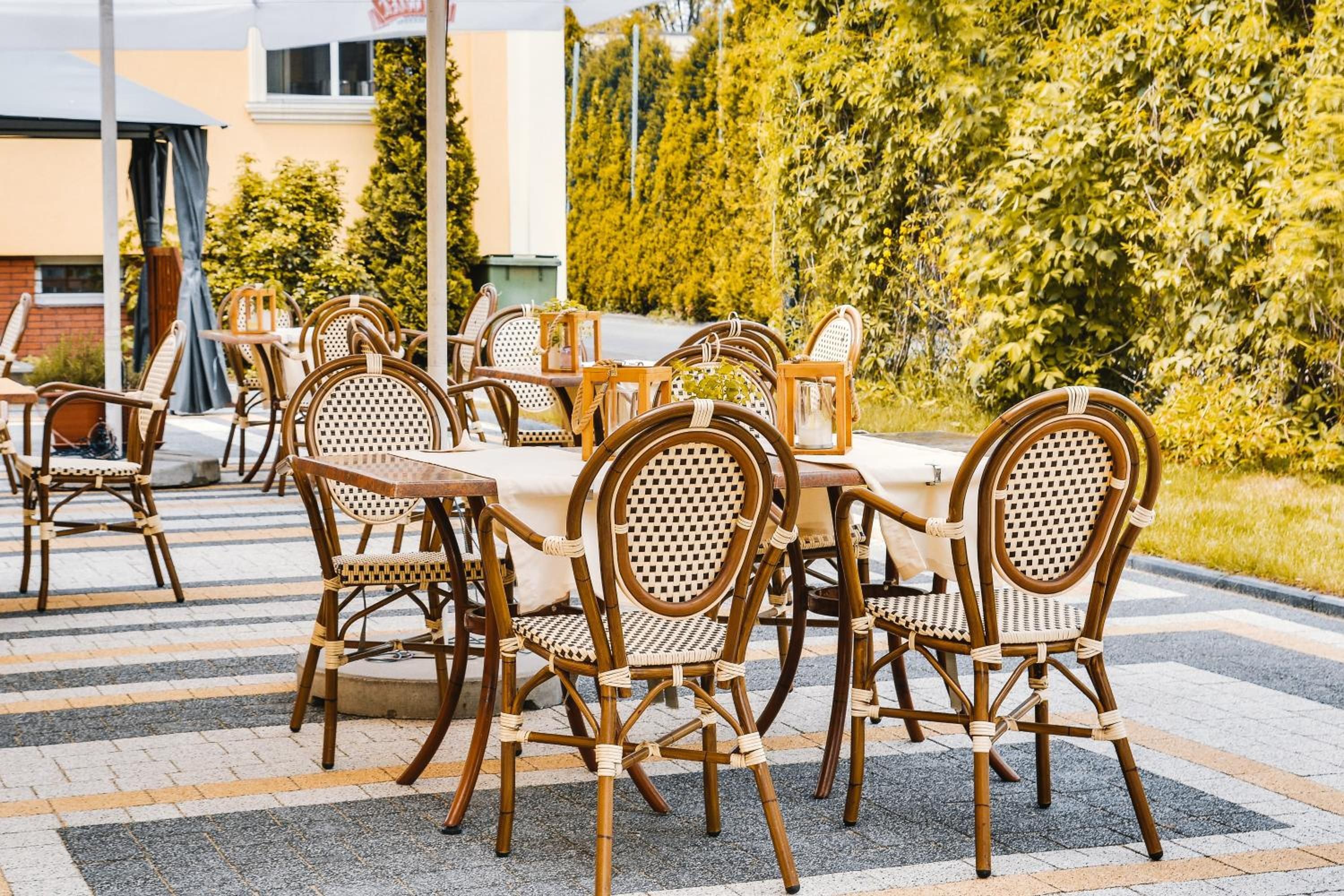 An outdoor dining area with wicker chairs and tables, surrounded by greenery and a building in the background.