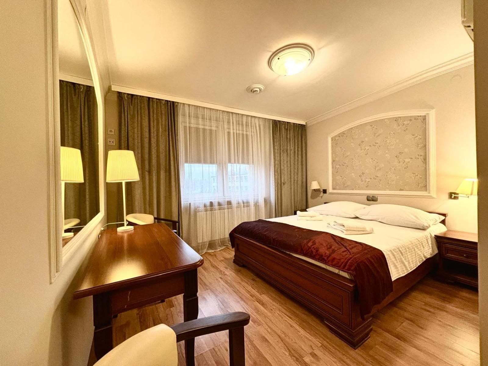 A hotel room with a wooden bed, a desk, a chair, a large mirror, a window with curtains, and a wall-mounted lamp.