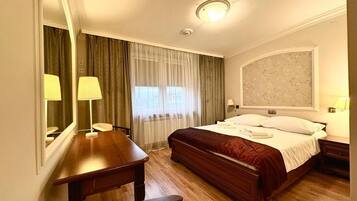 A hotel room with a wooden bed, a desk, a chair, a large mirror, a window with curtains, and a wall-mounted lamp.