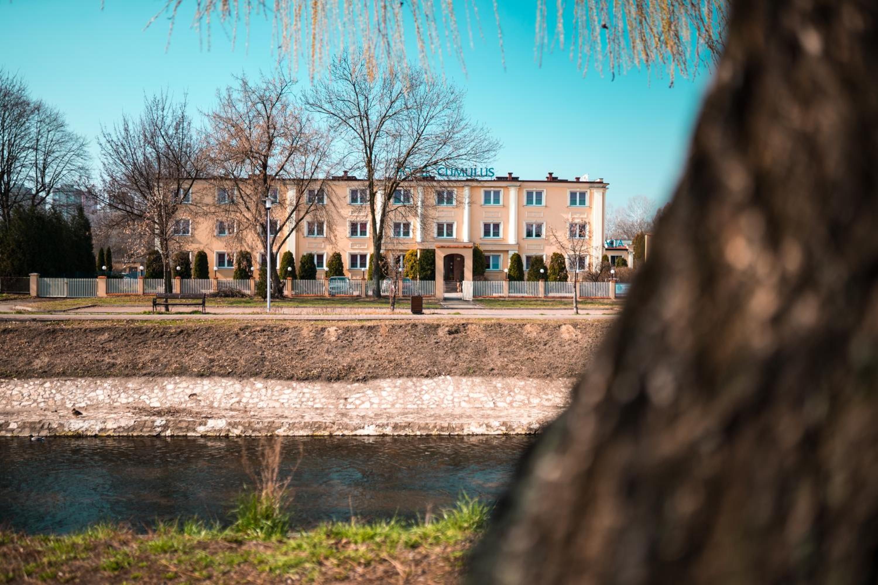 A multi-story hotel with a symmetrical facade, a paved courtyard, and a body of water in the foreground.