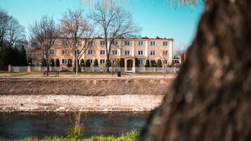 A multi-story hotel with a symmetrical facade, a paved courtyard, and a body of water in the foreground.