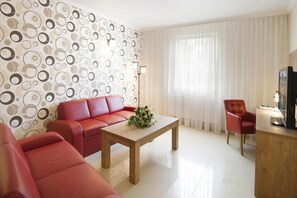 Una sala de estar moderna con un sofá rojo, una mesa de centro de madera y un sillón rojo. Se ve una pared decorativa con patrones circulares y un gran ventanal con cortinas translúcidas.