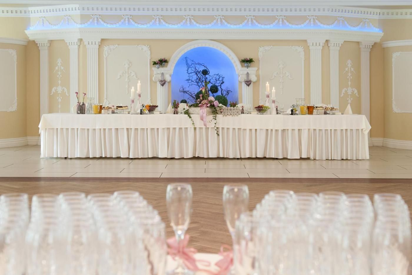 Une salle de banquet avec une longue table dressée pour un événement formel, flanquée de colonnes décoratives et d’une grande fenêtre en arc de cercle.