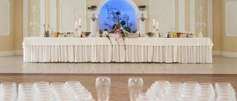 Une salle de banquet avec une longue table dressée pour un événement formel, flanquée de colonnes décoratives et d’une grande fenêtre en arc de cercle.