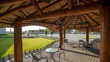 Un patio exterior con techo de paja, zona de estar y vistas a un jardín ajardinado y una carretera.