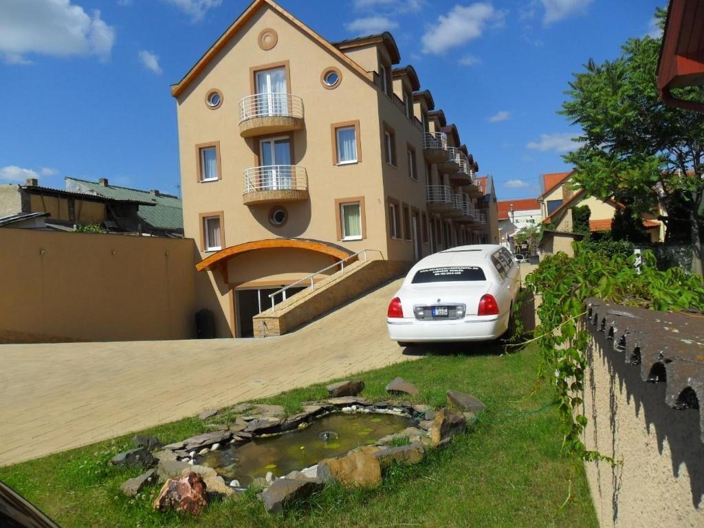 A multi-story building with balconies, a car parked in front, and a small pond with rocks.