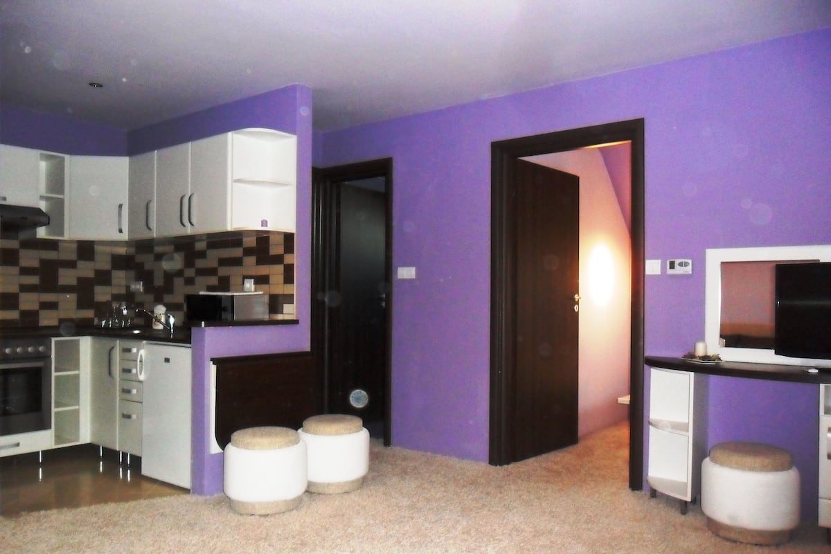 A compact kitchen with white cabinets, a microwave, and a small countertop. There are two stools and a door leading to another room.
