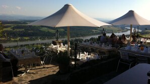 An outdoor dining area with tables and chairs, umbrellas, and a scenic view of a lake and hills.