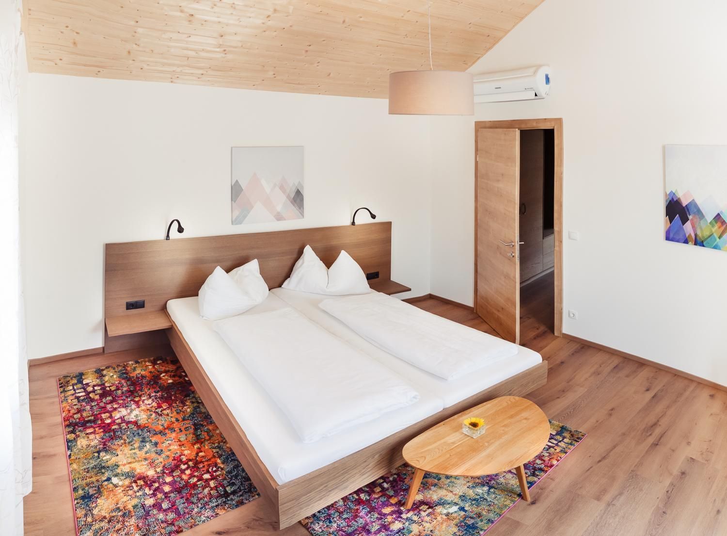 A modern bedroom with a wooden bed, a colorful rug, a small wooden table, and a door leading to another room.