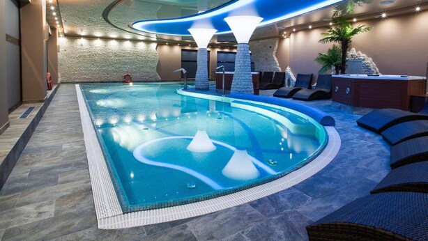A modern indoor swimming pool with a curved design, surrounded by lounge chairs and a stone wall. - Hotel Zawiercie (Zawiercie)