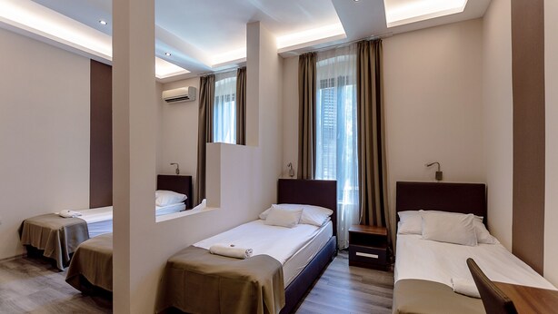 A hotel room with three single beds, a large mirror, an air conditioning unit, and a view of the city through the window. - Vén Diófa De Lux Panzió (Szeged)