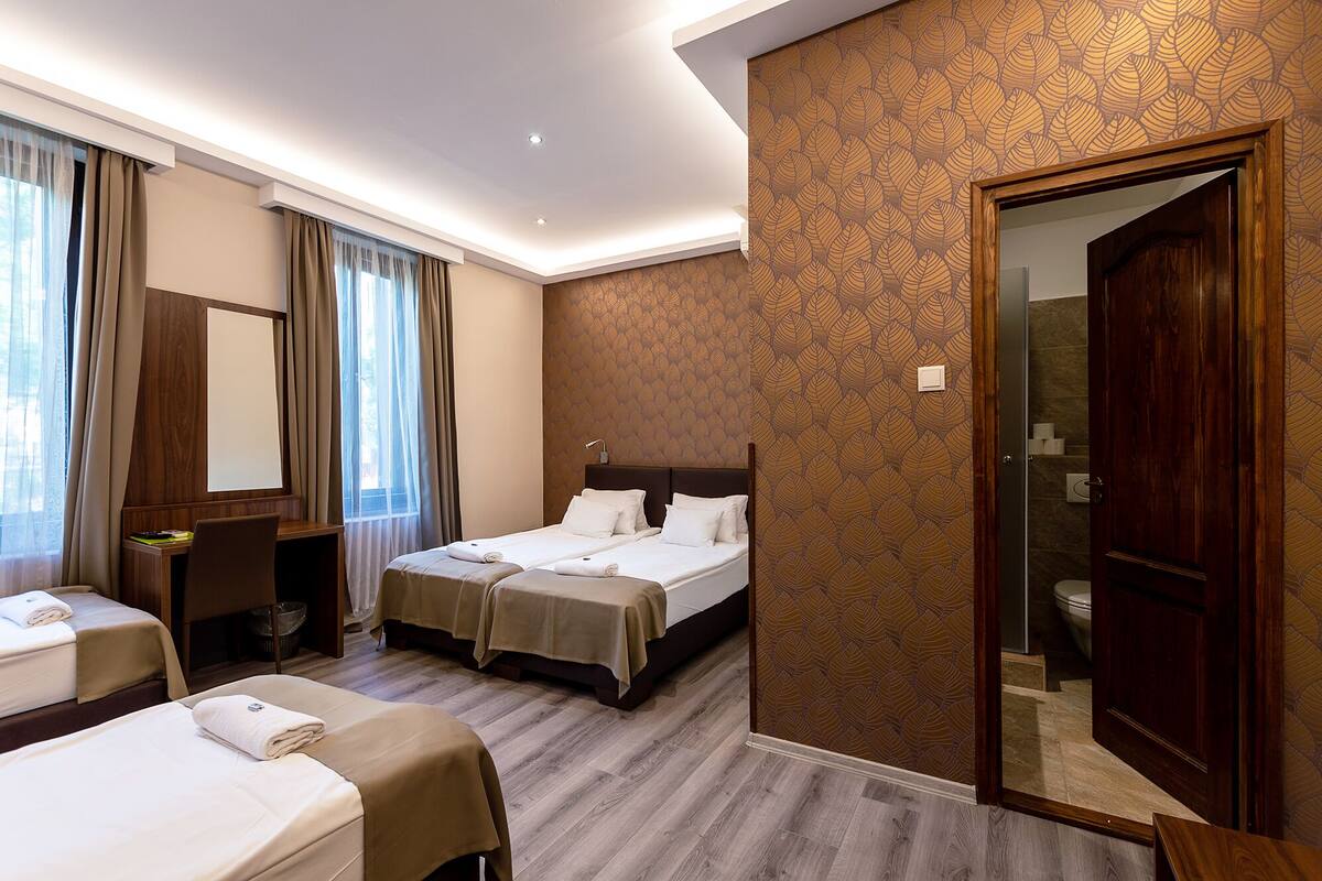 Habitación de hotel con dos camas, un escritorio, una silla, un espejo y un baño visible a través de una puerta abierta.