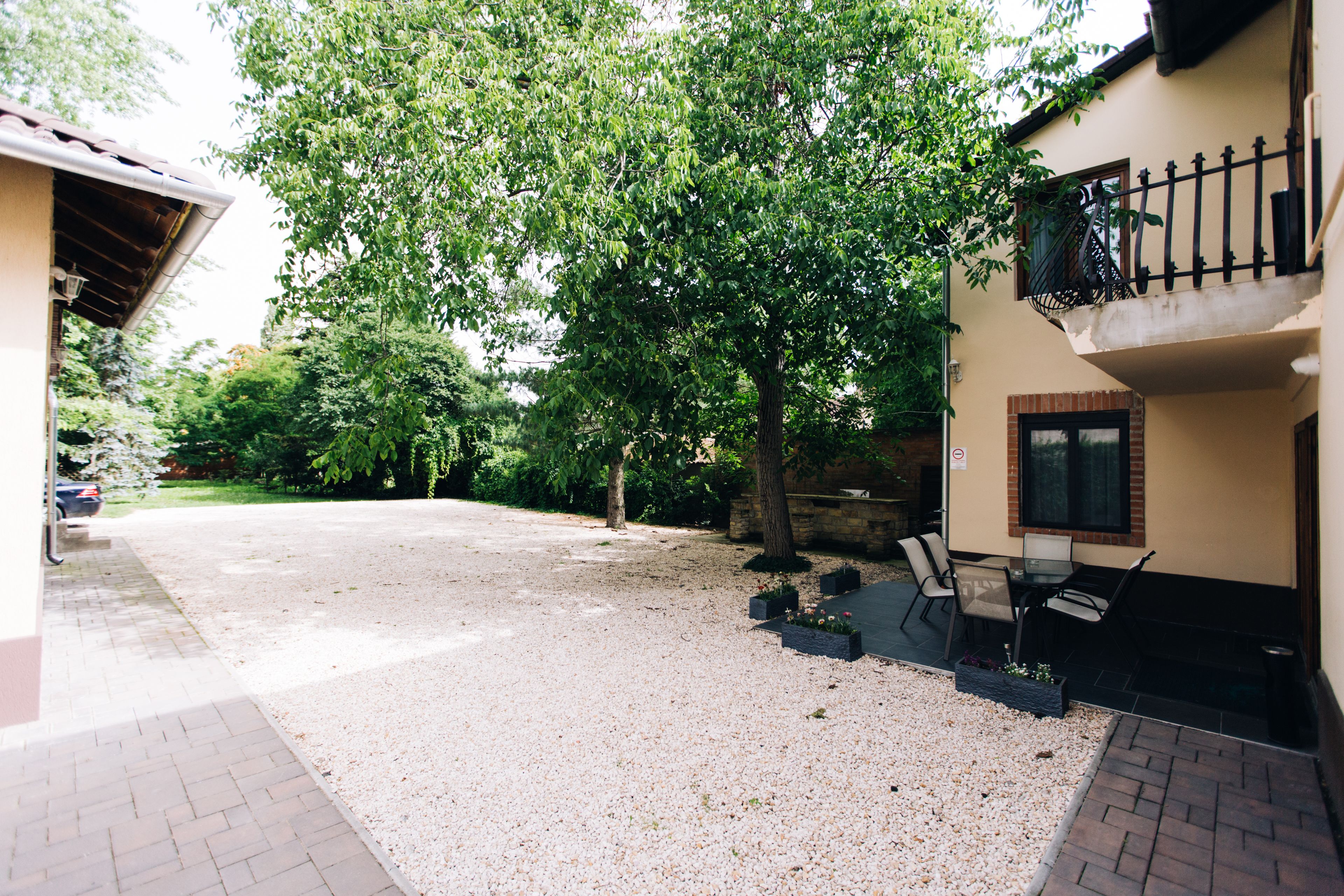 A residential area with a gravel driveway, a tree, a patio with outdoor furniture, and a balcony with a metal railing.