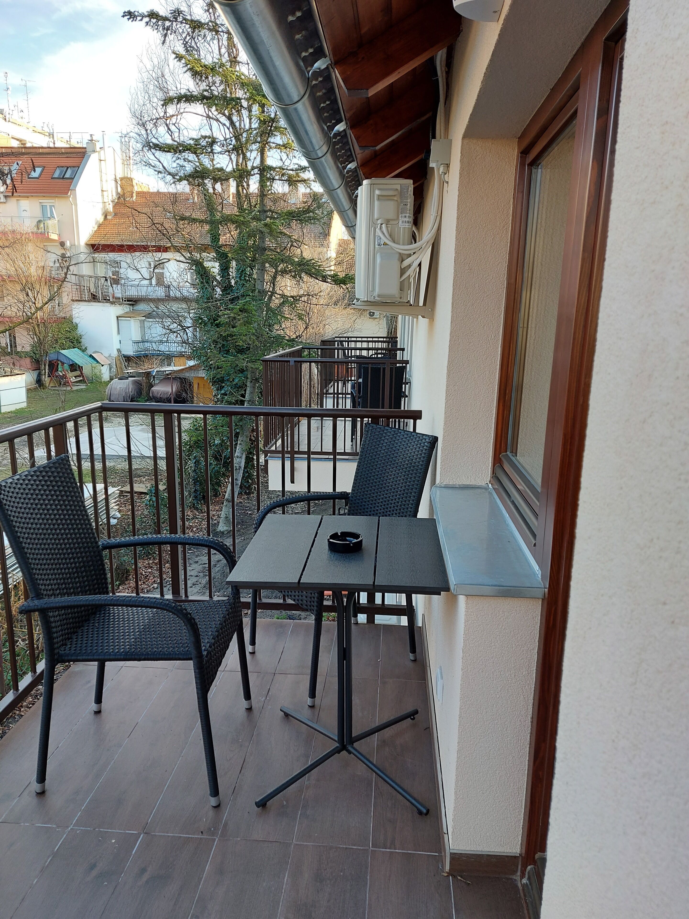 A balcony with a table and chairs, a view of the neighborhood, and a building with a window and air conditioning unit.