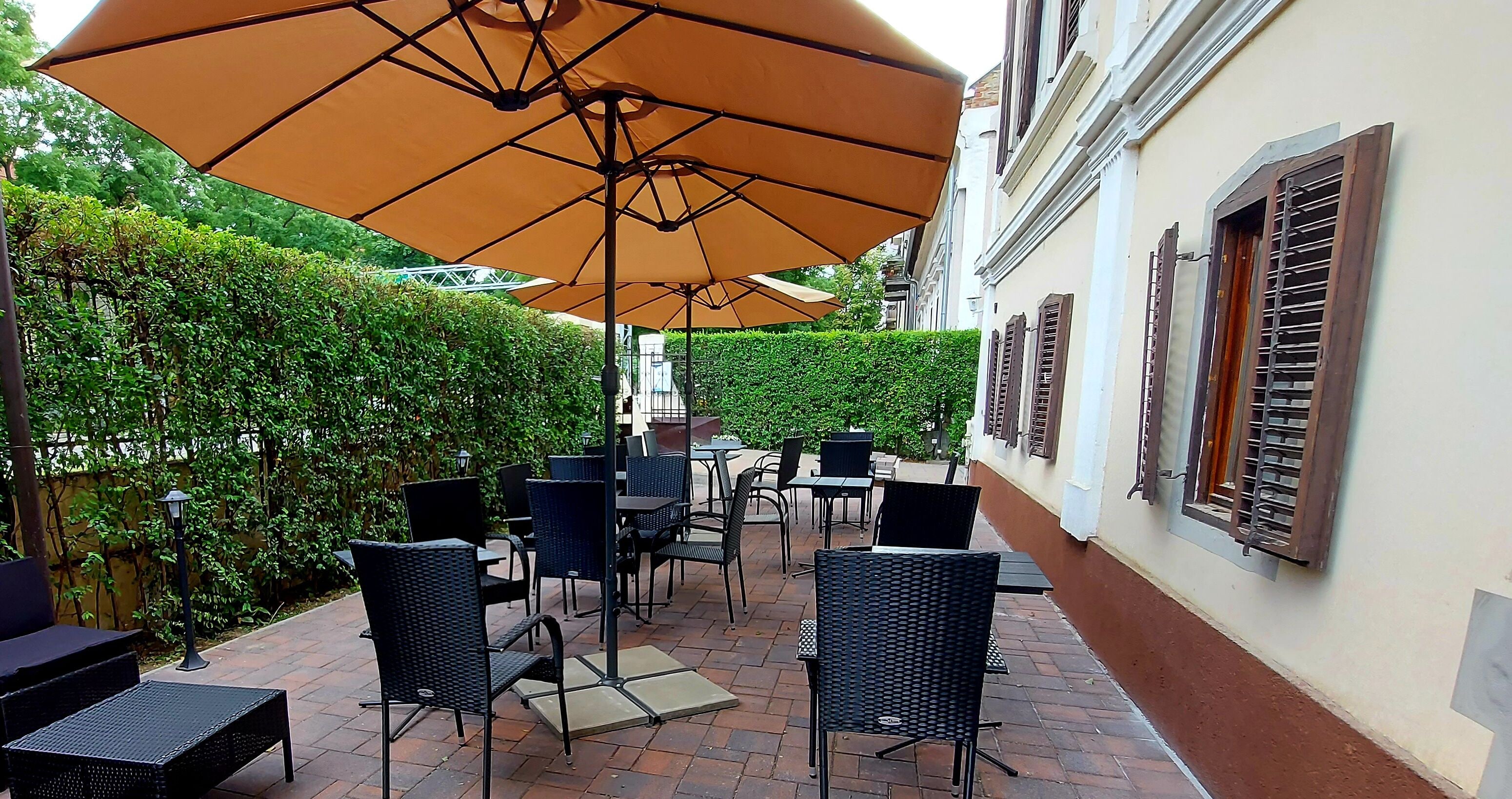 An outdoor patio with wicker furniture and large umbrellas, surrounded by a hedge and a building with shutters.