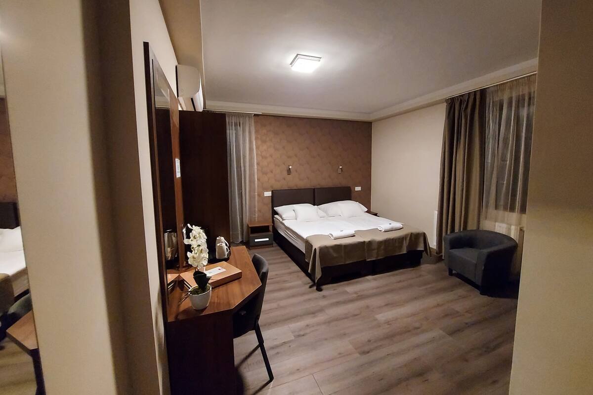 Habitación de hotel con cama, escritorio con silla, pequeña mesa con planta y ventana con cortinas.