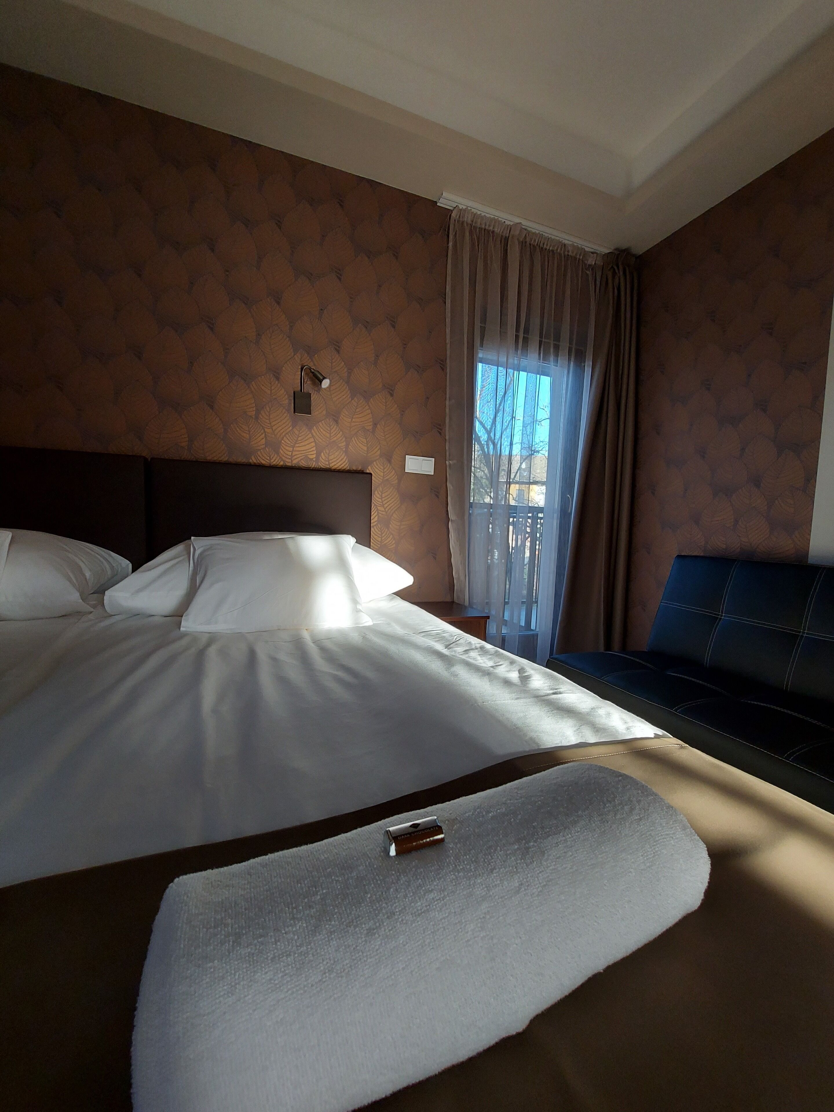 Hotellihuone, jossa on suuri sänky, yöpöytä, ikkuna verhoilla ja näkymä ulos.