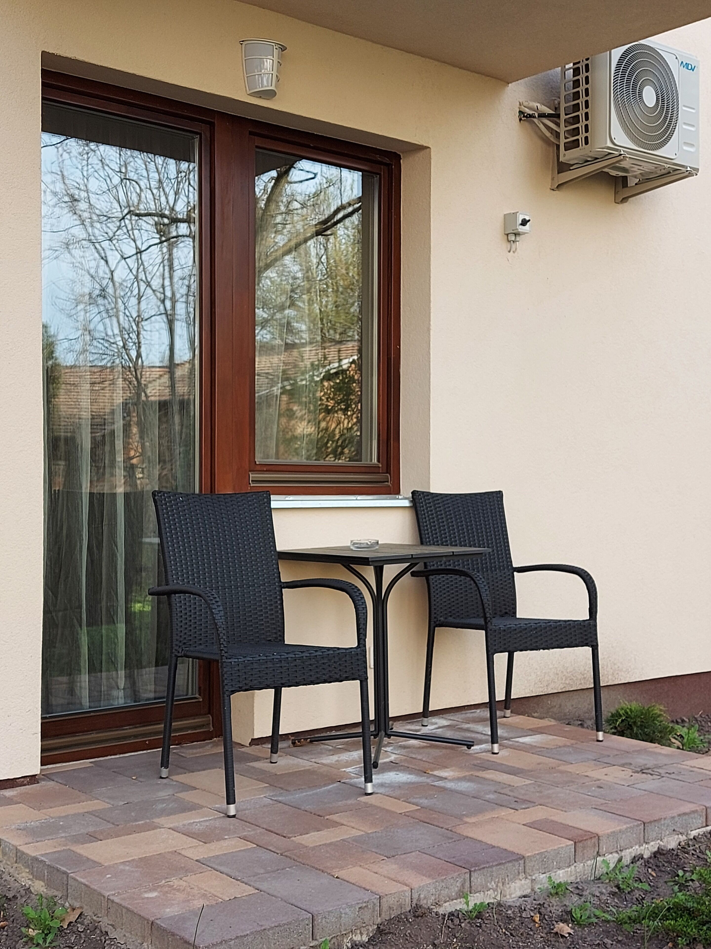 An outdoor patio with two wicker chairs and a small table, a window with a view of trees, and an air conditioning unit mounted on the wall.