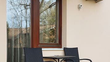 An outdoor patio with two wicker chairs and a small table, a window with a view of trees, and an air conditioning unit mounted on the wall.