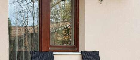 An outdoor patio with two wicker chairs and a small table, a window with a view of trees, and an air conditioning unit mounted on the wall.