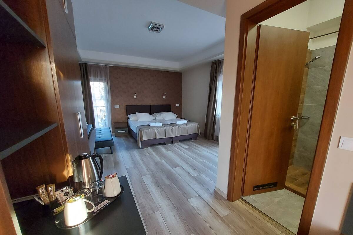 Habitación de hotel con cama, una mesita con hervidor y tazas, zona de ducha y puerta de madera.