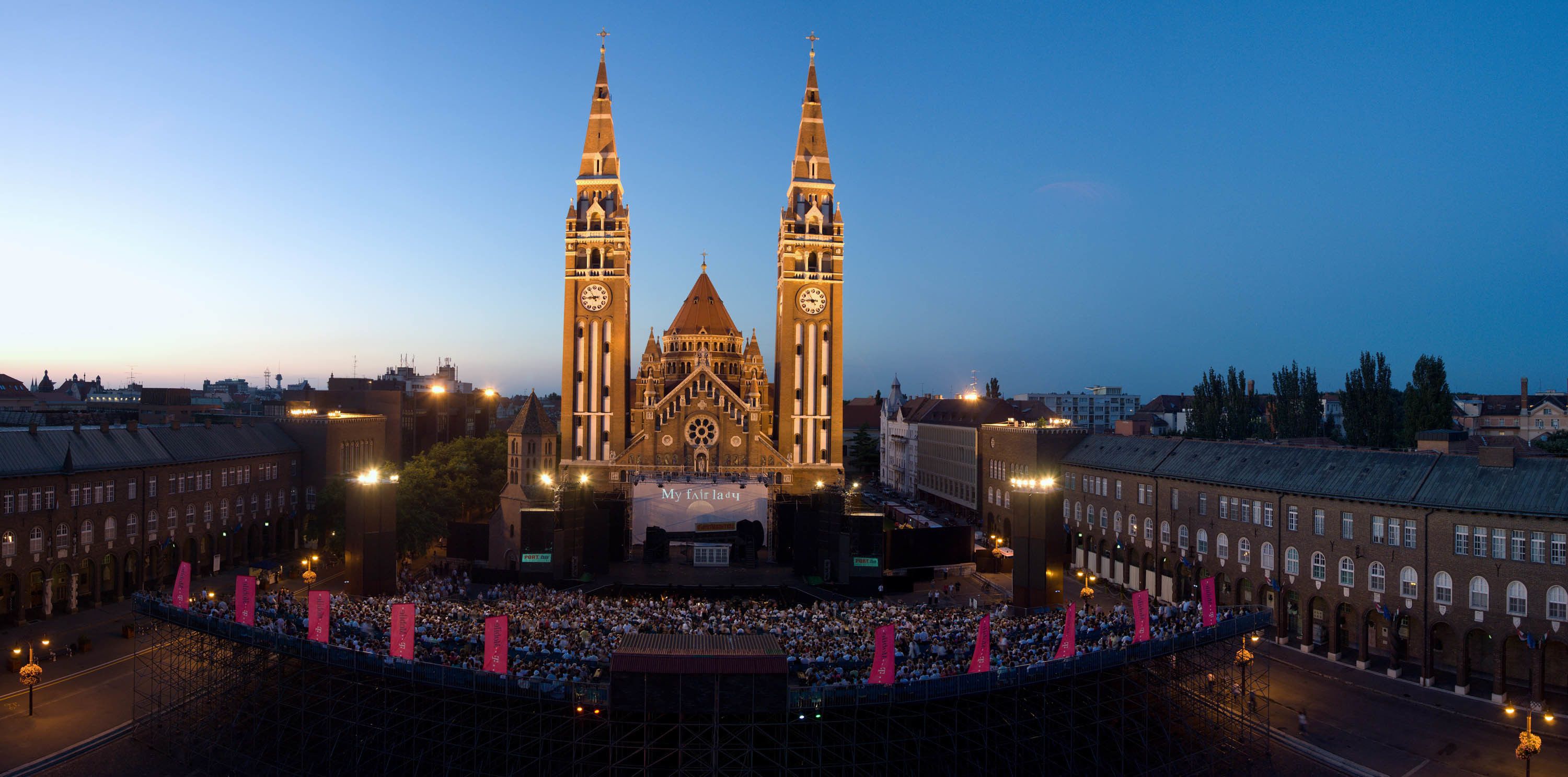 A historic building with two prominent towers, a large crowd gathered in front, and a stage set up for an event.