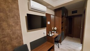 A modern room with a flat-screen TV mounted on the wall, a wooden desk with a chair, a small sofa, and an air conditioning unit. - Vén Diófa De Lux Panzió (Szeged)