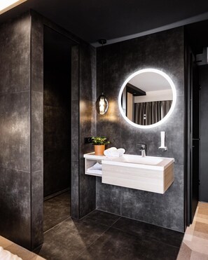 A modern bathroom with a dark wall, a round mirror with a light, a white vanity, and a potted plant.
