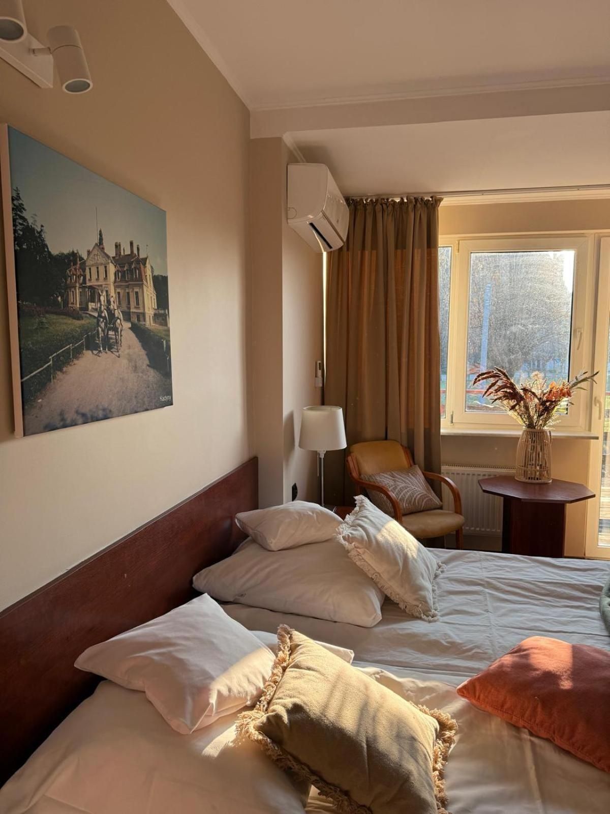 A hotel room with a bed, a chair, a lamp, a window with curtains, and a painting on the wall.