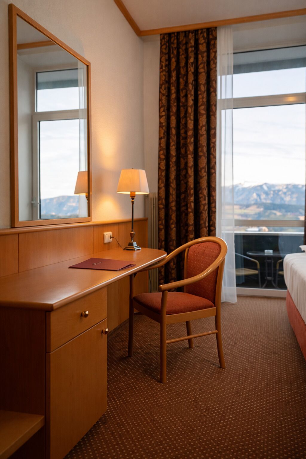 A hotel room with a wooden desk, a chair, a bed, a large window with a view, and a mirror.