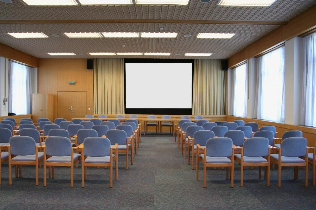 A conference room with rows of chairs facing a large screen, wooden panel walls, and large windows.