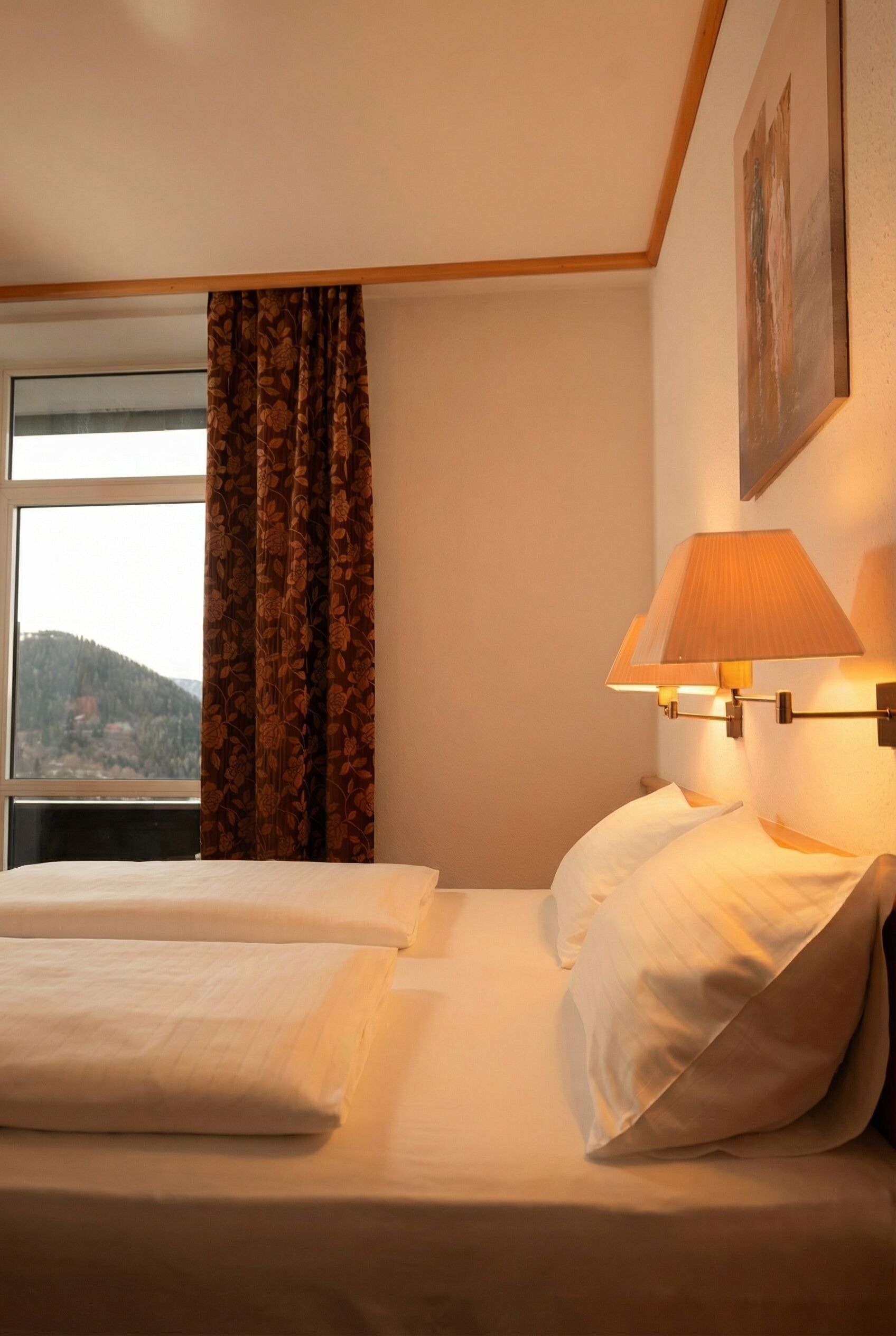 A hotel room with a bed, a window with curtains, a wall-mounted lamp, and a painting.