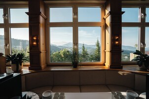 Quarto com uma janela ampla com vista para uma paisagem montanhosa, um espaço de estar com assentos acolchoados e uma mesa com pratos e xícaras.