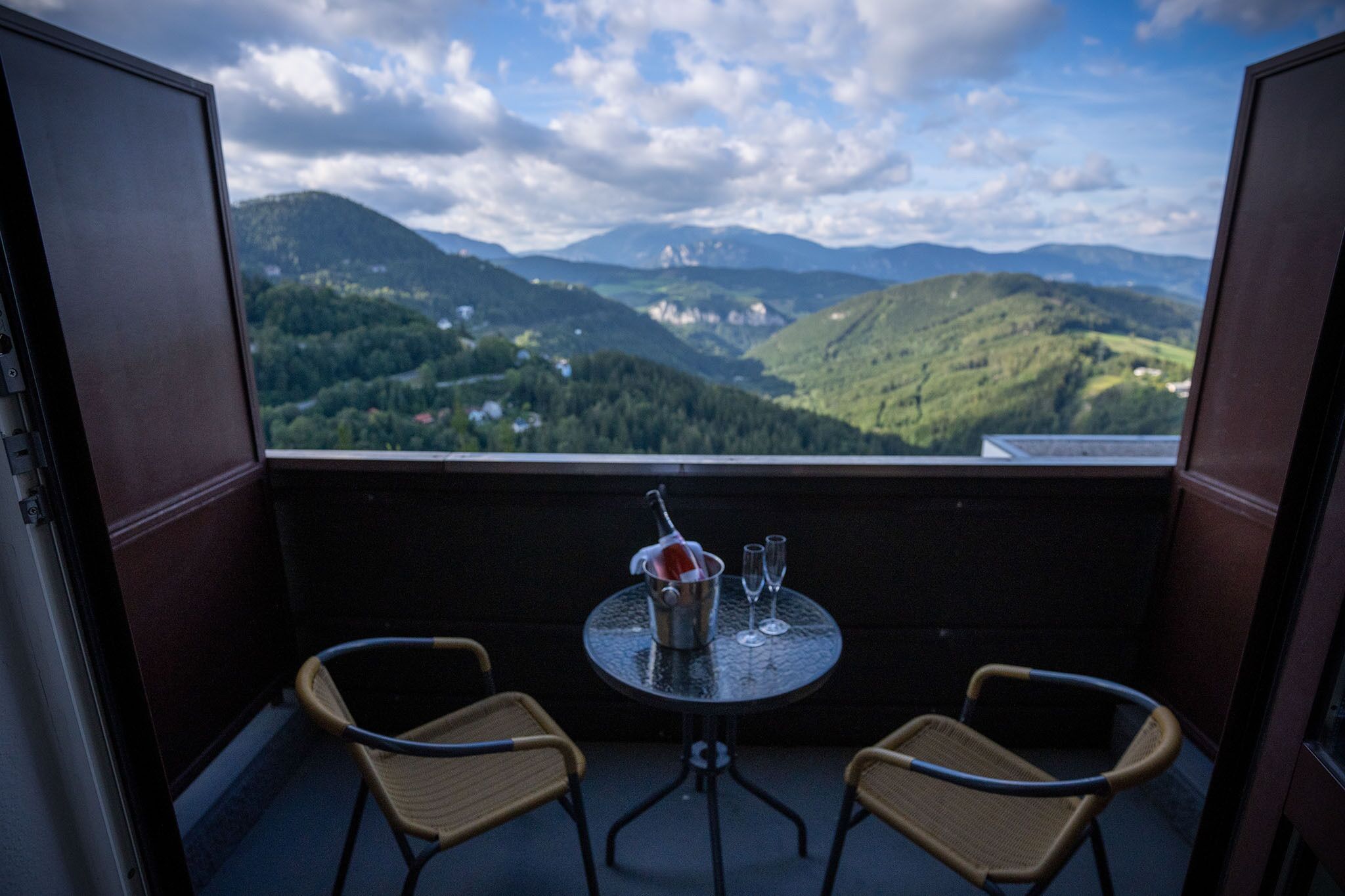 Ein Balkon mit zwei Korbstühlen, einem kleinen runden Tisch und einer Champagnerflasche. Der Balkon bietet Blick auf eine gebirgige Landschaft mit Bäumen und Gebäuden in der Ferne.