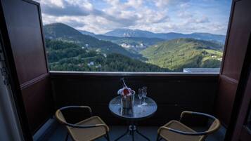 Ein Balkon mit zwei Korbstühlen, einem kleinen runden Tisch und einer Champagnerflasche. Der Balkon bietet Blick auf eine gebirgige Landschaft mit Bäumen und Gebäuden in der Ferne.