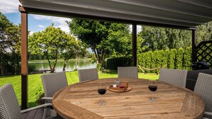 Un espacio de estar al aire libre cubierto, con una mesa de madera, sillas de mimbre y vistas a un lago y árboles.