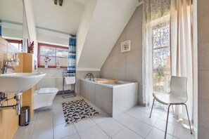 A modern bathroom with a large bathtub, a sink, and a toilet. There is a window with a view of the sky and a curtain.