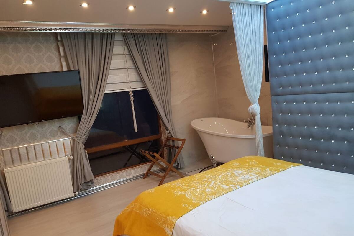 A bedroom with a bed, a bathtub, a radiator, a television, and curtains.