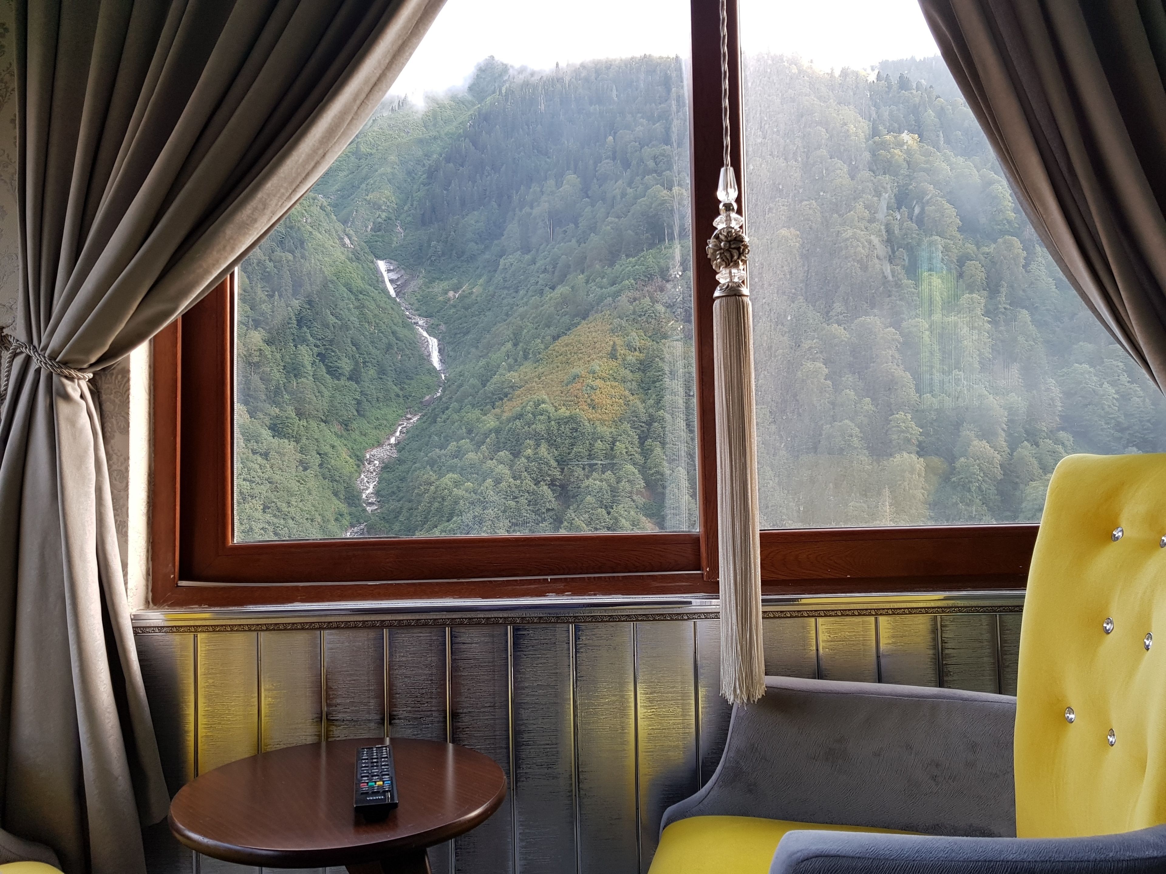 A room with a large window offering a view of a forested valley and a waterfall. There is a small table with a remote control on it, and a yellow armchair.