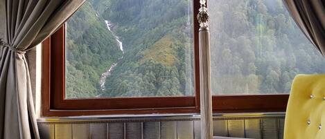 A room with a large window offering a view of a forested valley and a waterfall. There is a small table with a remote control on it, and a yellow armchair.