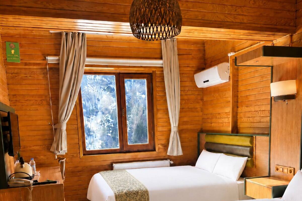 A wooden cabin room with two beds, a wooden dresser, a hanging light fixture, a window with curtains, and a wall-mounted heater.