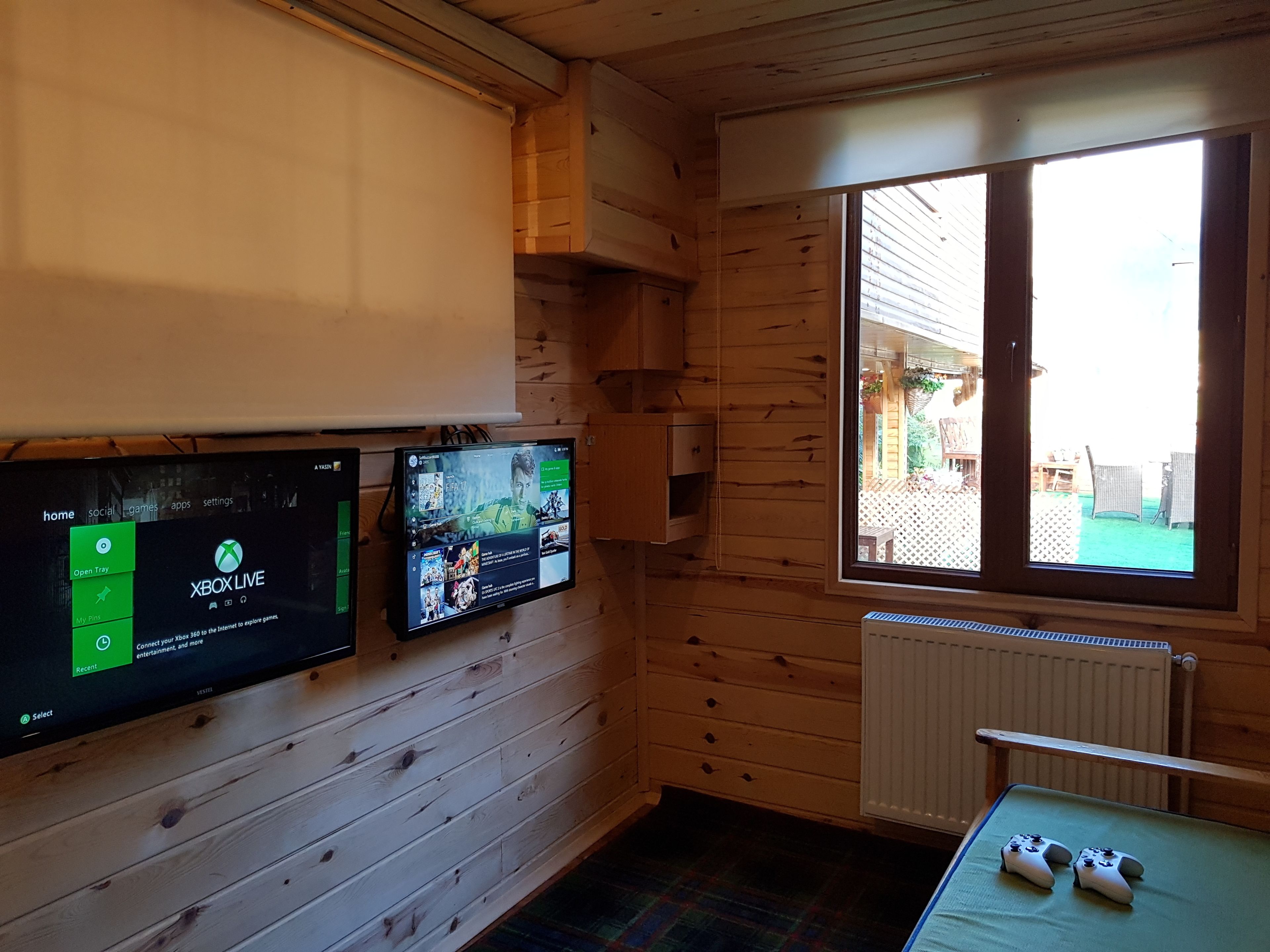 Een kamer met houten wanden, een groot raam en twee bevestigde schermen waarop Xbox Live te zien is.
