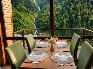 Una mesa de comedor con platos, cubiertos y un jarrón con flores, con el fondo de un valle boscoso y una cascada.