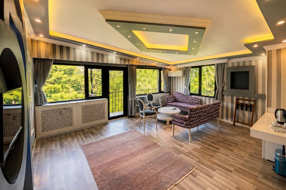 A modern living room with a sofa, chairs, a TV, and large windows with a view of greenery.