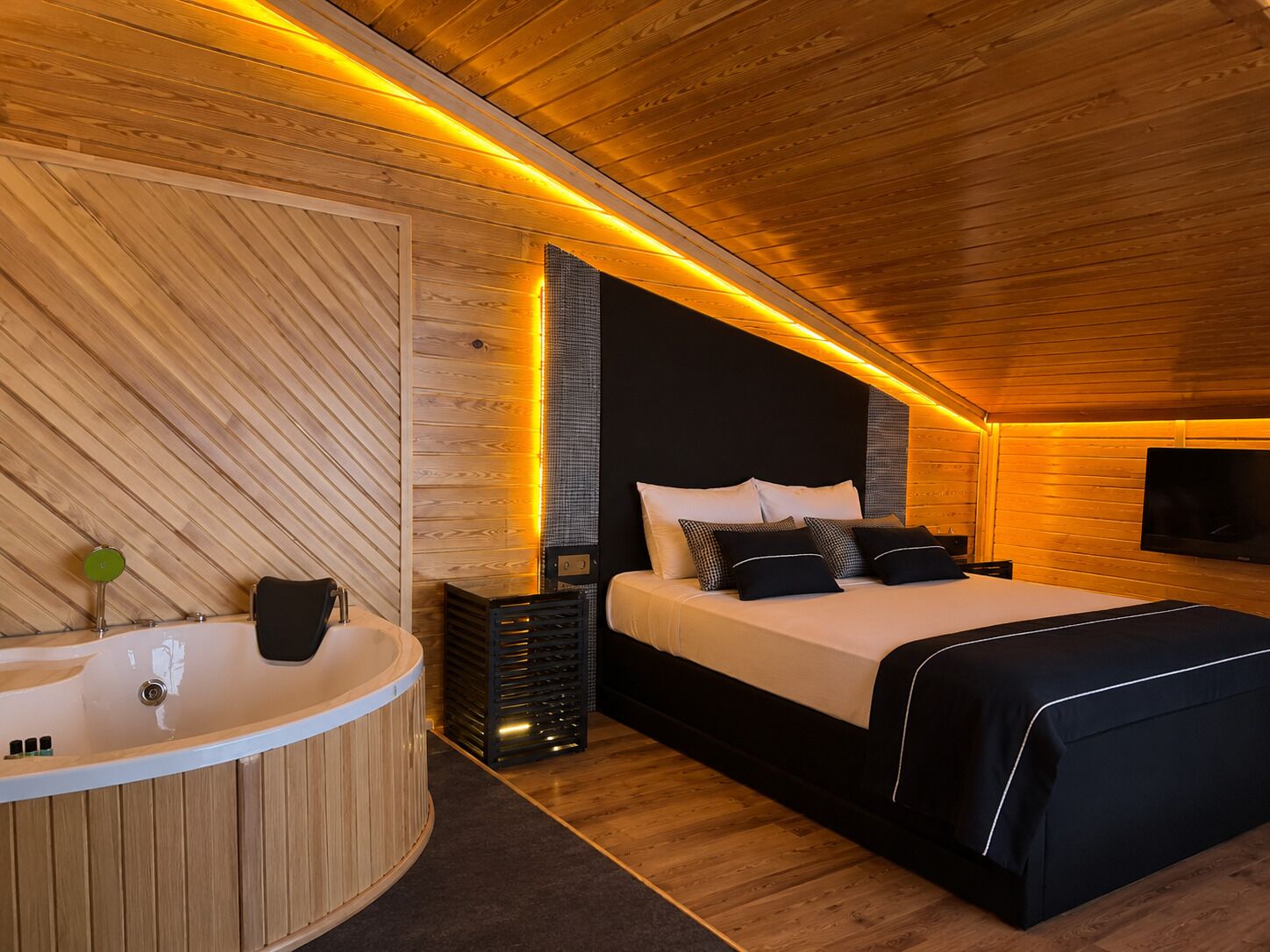 Ein modernes Schlafzimmer mit Holzboden, einem großen Bett mit schwarzer Bettwäsche, einem an der Wand montierten Fernseher und einer Holzwanne.