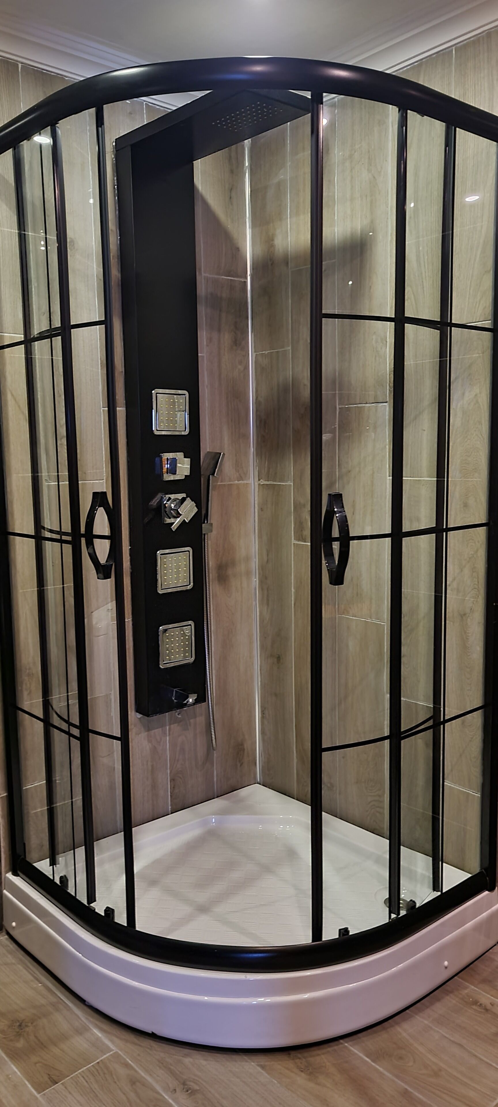 Un espace de douche d’angle doté d’une porte en verre, d’un panneau de commande et d’un sol carrelé de couleur blanche.