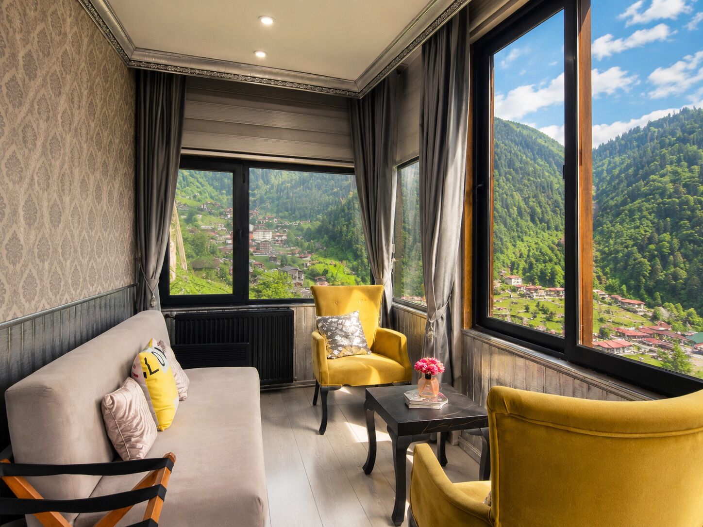 A room with a sofa, two armchairs, a table, and a view of a green valley through large windows.