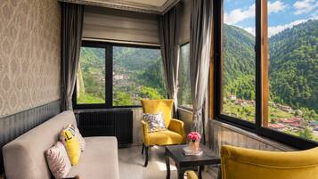 A room with a sofa, two armchairs, a table, and a view of a green valley through large windows.